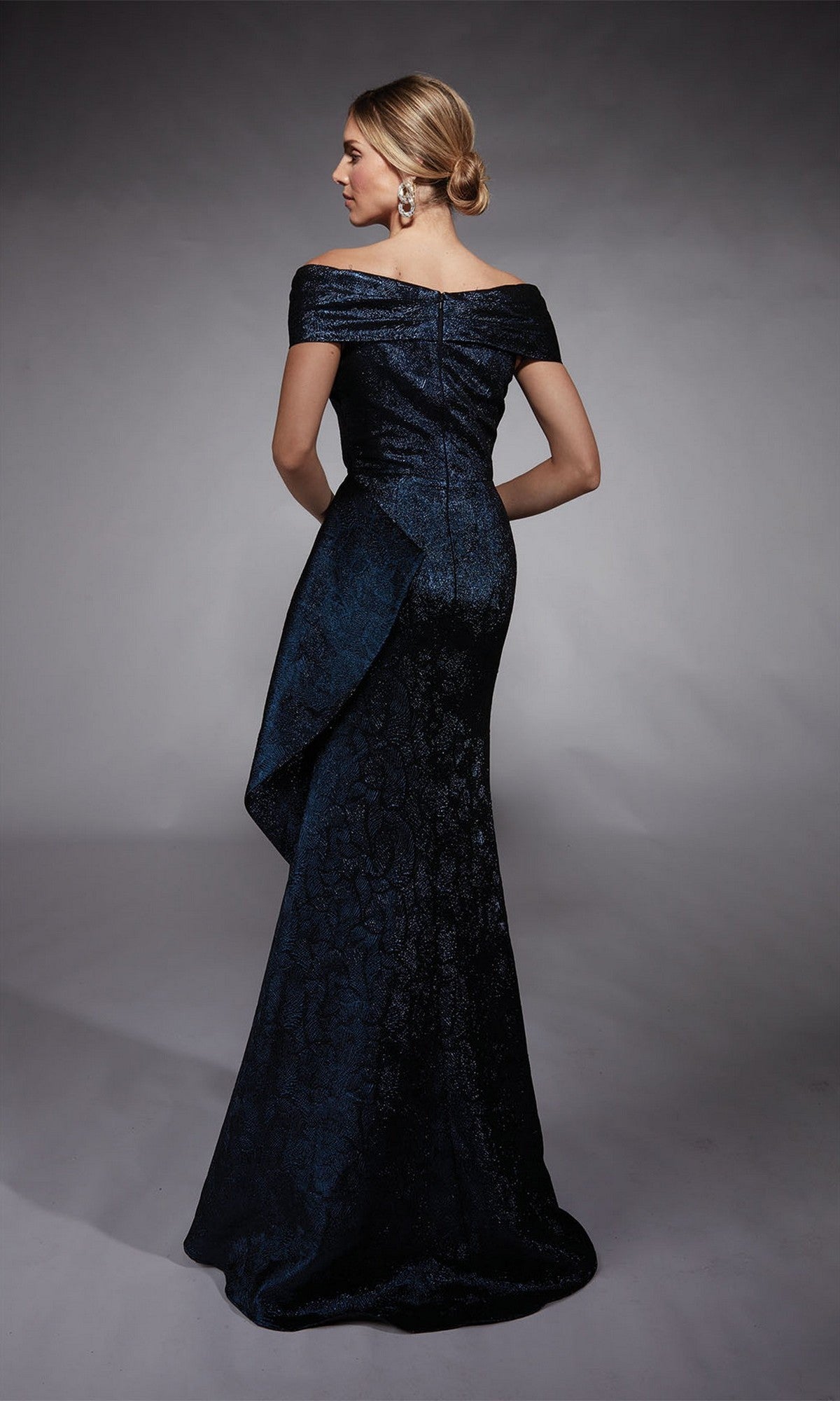 A woman with a low updo faces away, wearing the Alyce 27774 long formal off-the-shoulder mermaid gown in dark blue, featuring textured patterns and draped detailing.